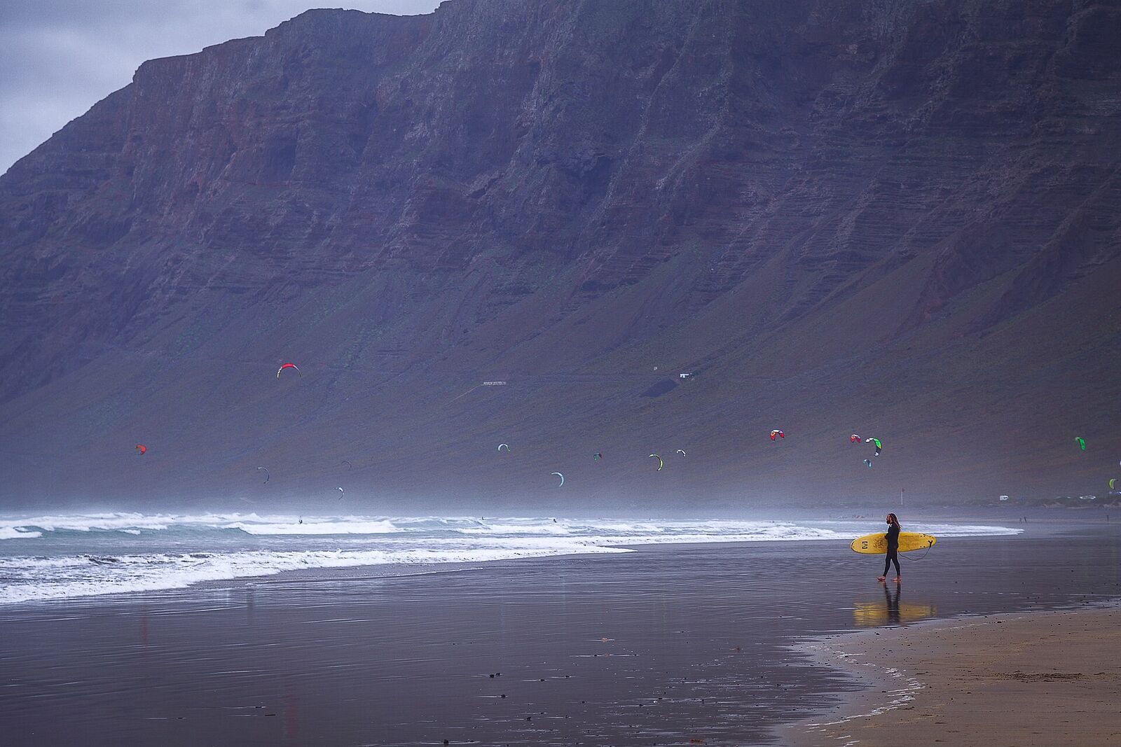 A surfer walking the waterline at Famara beneath the Risco de Famara cliffs, with kitesurfers out in the bay